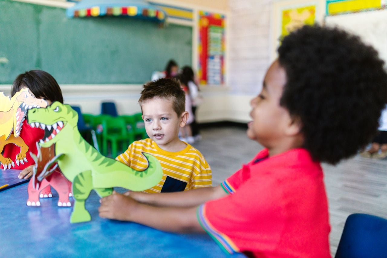 Crianças de 5 anos brincando juntas com dinossauros de papel em volta da mesa da sala de aula